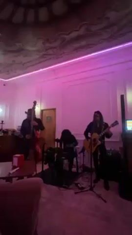 A live band is performing in a room with pink lighting. The band consists of three members: one playing a double bass, one playing a small drum kit, and one playing an acoustic guitar while singing. The room has white walls with decorative molding and an ornate ceiling. There are tables and chairs visible in the foreground, suggesting the band is playing in a bar or restaurant setting. There is also a closed door to the side and an open area behind the bassist. A speaker is seen behind the guitarist.