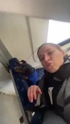 A woman on a train takes a selfie with a small black and tan dog wearing a blue harness sitting on the seat behind her. The train car is mostly empty, with blue seats and large windows. Outside the window, there is snow on the ground and bare trees. The train car is lit by fluorescent lights.