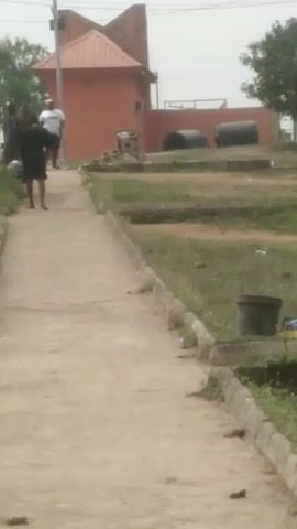 The scene takes place on a hazy day, in what appears to be a park or outdoor recreational area. The focal point is a concrete pathway leading towards a small, orange-colored building with a pitched roof. Adjacent to the path are grassy areas with concrete borders that resemble garden beds.  There are also large black cylindrical containers visible in the background.

Several people are present in the video. Initially, three individuals are standing near the building, but they are somewhat blurred. Later, three women, dressed in casual attire, walk along the path towards the building. Their clothing includes shorts and tank tops. In addition to the women, the three individuals are joined by a man in a white shirt.

The overall setting appears to be somewhat unkempt, with overgrown grass and some discarded trash visible. The camera remains stationary throughout the short clip.
