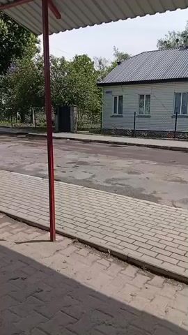 The video shows a blue bus driving on an asphalt road. The video is taken from under a small bus stop. The road is flanked by brick walkways and simple homes with some greenery in the background.