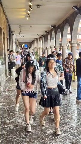 The video shows a group of young people participating in what appears to be a festive event. They are walking and laughing in an open arcade with stone arches and columns. They are covered in white foam. The ground is wet with the foam. The group consists of both young men and women, wearing casual clothing such as shorts, skirts, t-shirts, and light jackets. Some are smiling and playfully wiping the foam off their faces and clothes. Beyond the arcade, there is a glimpse of an open square filled with more people, suggesting a larger celebration or festival is taking place. The atmosphere seems joyful and carefree.