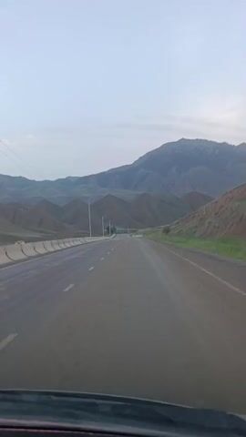 This media file captures a point-of-view shot from a vehicle traveling along a paved road. The road stretches out towards a horizon dominated by rolling, arid mountains. The sky is a pale, overcast blue, suggesting an early morning or late afternoon setting with diffused light. The weather appears to be mild, with no visible precipitation or strong sunlight.

The scene is characterized by the vast, natural landscape. The mountains are a prominent feature, displaying various shades of brown and green, with some rocky outcrops visible on the higher peaks. The road itself is a two-lane highway with a concrete barrier on the left side and a shoulder on the right. White dashed lines mark the lanes. Several utility poles with wires run alongside the road, indicating infrastructure in this somewhat remote area.

There are no people or animals visible in the frame. The primary "object" is the road and the surrounding natural environment. The activity is clearly travel, with the perspective suggesting the vehicle is moving forward. The absence of harsh shadows and the muted colors contribute to a calm and perhaps slightly melancholic or contemplative mood. The location cues, such as the type of mountains and the sparse vegetation, are consistent with the Naryn region of Kyrgyzstan. No text is visible in the media file.