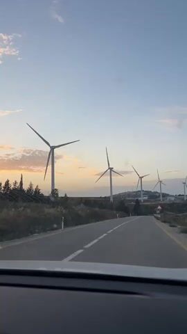 The video shows a view from inside a car driving on a paved road towards a row of wind turbines. The sky is a pale blue with some light orange clouds near the horizon, suggesting it is either sunrise or sunset. The road is asphalt with a white line down the center. On either side of the road is dry scrub vegetation. The wind turbines are tall and white, with three blades each. The camera appears to be stationary inside the car, with minimal movement or zooming. There are no people visible in the video.