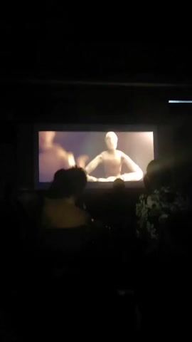 A group of people are watching a projection on a screen in a dark room. The projection displays a wooden mannequin with its arms outstretched, sitting at a table. The mannequin is illuminated by a bright light, and there are blurred figures in the background of the projection. The people in the foreground are silhouetted against the screen and are seated. One person in the foreground wears a yellow shirt, another a patterned shirt, and a third person's clothing is indistinct. The room appears to be a theater or screening room.