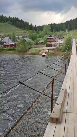The scene is captured from what appears to be a bridge, offering a view of a river flowing swiftly. The water is dark and textured, reflecting the overcast sky. On the opposite bank, a small village nestles at the base of a green hillside covered with trees. Several houses with varying colors of roofs can be seen, some with bright accents such as red or blue. Along the riverbank, kayaks are visible. The bridge itself has a chain-link fence and wooden planks.