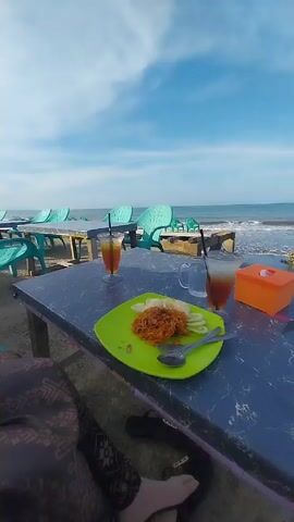 The video shows a green plate of food and two glasses of iced tea on a dark blue table. The plate contains what appears to be fried noodles, sliced cucumbers, and several white crackers. There are also a fork and spoon on the plate. One glass of iced tea is mostly finished, with the other still full. Beside the glasses is a small empty glass mug and a small orange tissue box. The table is located at a beachside restaurant with blue plastic chairs visible in the background. The sea and a clear blue sky are also visible in the background. In the foreground, parts of a person's patterned pants and black sandals can be seen. A soft hum is audible in the audio.