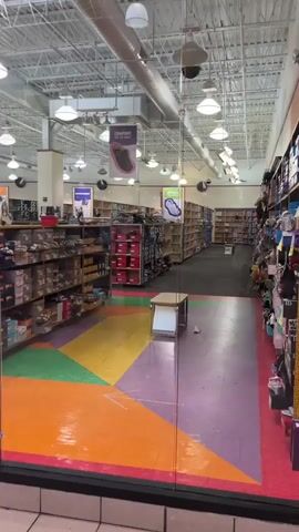 The video pans across the storefront of a shoe store in a mall. The floor inside the store is a colorful geometric pattern of orange, yellow, green, and purple. Shelves filled with shoe boxes line the walls. Several backpacks are displayed on a rack near the entrance. The store appears to be empty of customers and employees.
