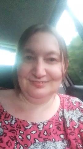 This media file features a medium close-up shot of a woman in what appears to be a car, likely Berezynka, Ukraine, given the context. The woman is smiling and looking towards the camera. She has light brown hair and is wearing a vibrant pink t-shirt with a repeating pattern of white and black leopards. The lighting suggests it might be daytime, possibly overcast or late afternoon, as the light is somewhat diffused. The background is blurry, showing hints of trees and the interior of a vehicle, but provides no specific details about the location. The woman's expression is pleasant and relaxed, suggesting a casual or friendly interaction. There is no visible text in the frame.