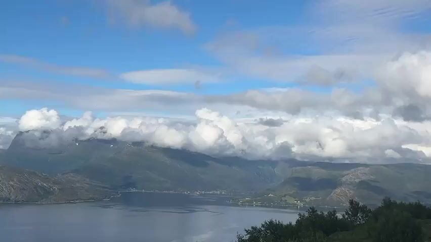 A panoramic video captures a vast body of water framed by rugged, mountainous terrain under a bright, partly cloudy sky. The water, appearing calm and deep blue, reflects the sky and surrounding landscape. The mountains, a mix of rocky outcrops and sparsely vegetated slopes, rise dramatically from the water's edge, some peaks partially obscured by fluffy white clouds that drift across the scene. The sky is a vivid blue, streaked with wispy cirrus clouds and dotted with more substantial cumulus formations. The overall impression is one of serene natural grandeur.