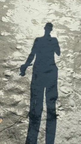 The video opens with a shadow of a person holding a fishing rod on a sandy beach. The camera then pans to reveal a wide, turquoise river flowing between lush green mountains. The riverbank is sandy, with some rocks and vegetation. The sky is clear and blue, with a few wispy clouds. The scene is peaceful and serene.