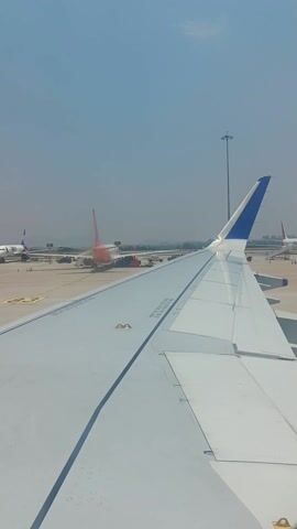 The video captures a bright daytime view from the window of an airplane on an airport tarmac. The prominent subject is the white wing of the aircraft in the foreground, with a distinctive blue tip on the far end, indicating an Indigo airline plane. Text "DO NOT WALK ON WING" is visible on the wing's surface.

In the background, several other airplanes are visible on the concrete tarmac. One particularly noticeable aircraft has a white fuselage and a vibrant orange/red tail, characteristic of SpiceJet airlines. Various airport ground equipment and vehicles are scattered around these planes. A tall light pole stands against the clear, blue sky on the right side of the frame. The weather appears sunny and clear.

An audible announcement plays, stating, "Ladies and gentlemen, boys and girls, we extend a warm welcome to our Indigo..." The video ends before the announcement concludes. The scene suggests the plane is either stationary at a gate, preparing for departure, or has just arrived.