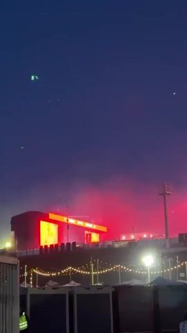 A low-angle, vertical shot captures a brightly lit outdoor stage under a twilight sky with a few green laser dots appearing in the top of the frame. The stage, viewed from a distance, features a large screen that changes colors from purple to red and displays text, though the specific content is not discernible. Below the stage, a row of booths or structures are illuminated by string lights, adding a festive ambiance to the scene. One person is visible in the bottom left, wearing a yellow safety vest. Smoke or fog emanates from the stage, suggesting a performance or special effects. The overall atmosphere suggests an evening event or concert.