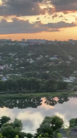This media file captures a picturesque sunset over the town of Rezina, Moldova. The scene overlooks a calm river, likely the Dniester, with its surface reflecting the warm hues of the sky and the surrounding greenery. On the far bank, a lush, tree-covered hillside rises, dotted with houses and a few larger buildings, including what appears to be a power plant or industrial facility with a tall smokestack.

The sky is a canvas of dramatic clouds, tinged with oranges, pinks, and yellows, indicating the sun is setting or has recently set. The light casts a warm glow over the landscape, creating a serene and tranquil atmosphere. The trees along the riverbank and on the hillside are full and green, suggesting it is late spring or summer.

The activity taking place is simply the observation of nature's beauty during sunset. There are no people or prominent objects visible in the foreground or mid-ground that detract from the natural scenery. The overall impression is one of peace and natural splendor. The time of day is clearly evening, as evidenced by the sunset colors and the soft, diffused light. The weather appears calm, with no strong winds evident in the reflections on the water. The location is confirmed as Rezina, Moldova, with the recognizable industrial structure on the hilltop serving as a key landmark. There is no visible text within the media file.