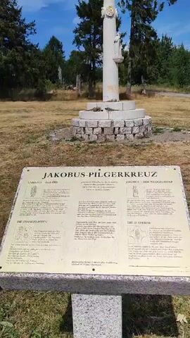 The video shows a close-up view of a sign in front of a stone cross. The sign has a gold-colored surface with text and illustrations. The title of the sign is "JAKOBUS-PILGERKREUZ" which translates to "Jacob's Pilgrim Cross". The sign is divided into several sections, including "CHRISTUS - DAS ZIEL" (Christ - The Goal), "DIE EVANGELISTEN" (The Evangelists), "JAKOBUS, PILGER WEGWEISER DURCH DAS JAHRLICHE GEBET, DEN GLAUBEN LEBEN" (Jacob, Pilgrim Guide through Daily Prayer, Living the Faith), and "DIE 12 STERNE" (The 12 Stars). The text and illustrations are in German. The sign is mounted on a stone base. In the background, the stone cross is visible, with a statue at the top. The cross is surrounded by a circular stone base. The area around the cross and the sign is grassy and dry, with trees in the distance under a blue sky with white clouds. The video pans slightly from left to right and then back, providing a clear view of the sign and the cross.