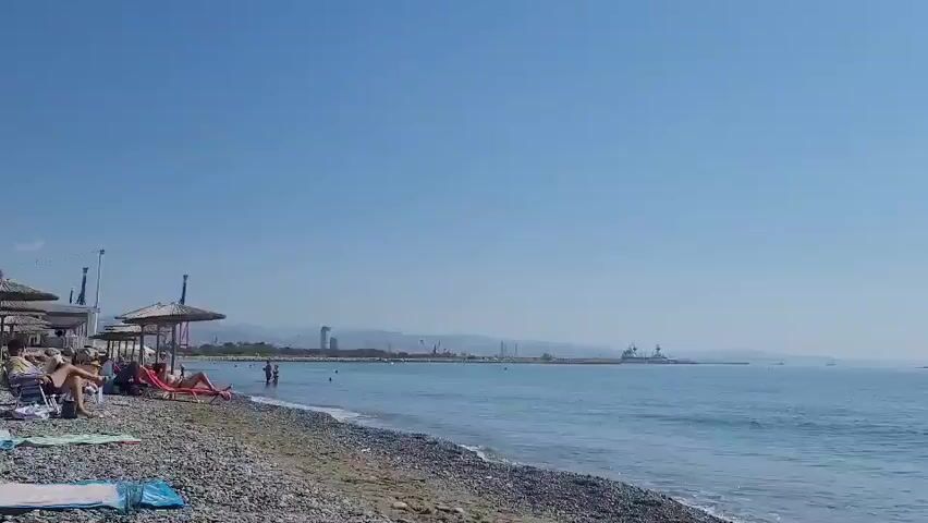 A sunny day at a pebble beach. The ocean is calm with gentle waves. Several people are visible on the beach, some under umbrellas, others sunbathing or swimming. In the distance, a harbor with cranes and buildings can be seen. The sky is clear and blue.