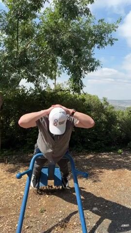 A man is performing back extensions on an outdoor fitness apparatus in Misgav Regional Council, Israel. He is wearing a grey short-sleeved collared shirt, dark pants, and a white baseball cap with three colored circles. His hands are behind his head. He bends forward at the waist, lowering his head towards the equipment, then returns to an upright position. The background features lush green trees, dense foliage, and a distant hilly landscape under a bright, cloudy sky, suggesting it is daytime. The fitness equipment is blue and metal with handles. The ground is covered in loose gravel and dirt. The overall scene is one of an outdoor workout in a natural, sunny environment.