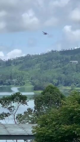 A seaplane flies across a cloudy sky over a green, hilly landscape. The plane is small and appears to be red and white. The landscape is covered in lush green trees, and a body of water is visible in the foreground. The sky is partly cloudy, with white clouds scattered across a blue sky. The plane appears to be in flight, moving from left to right across the frame.