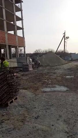 The video shows a construction site. A tall, unfinished building with exposed concrete and brick is visible on the left. In the foreground, there's a stack of rebar cages on wooden pallets, and piles of additional rebar laying nearby. A man in a yellow safety vest and work clothes is actively engaged in using ropes to maneuver rebar around the site, moving and adjusting the rebar and other bundles. Behind the rebar, there's a corrugated metal shed, a blue barrel, and other miscellaneous items. In the background, there's a large pile of sand or gravel, a cement mixer, and another person in work clothes. A power line stands further in the background, with bare trees visible. The ground is muddy and littered with debris. The sky is overcast.