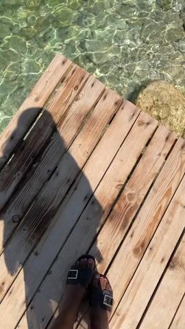 The video shows a close-up view of clear, shallow water with a rocky bottom. A wooden dock extends into the water from the lower left corner of the frame. A person's feet wearing black sandals are visible on the dock. Sunlight refracts through the water, creating shimmering patterns on the rocks below. The water is calm with gentle ripples. The scene conveys a sense of tranquility and a natural, pristine environment.