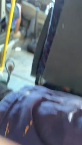 The video begins with a close-up view of a person's legs and feet, wearing dark socks and blue jeans, sitting inside a bus. A yellow pole is visible to the left. The camera then pans slightly to the right, revealing the interior of the bus, including a dashboard with a steering wheel and various controls. A person in the driver's seat is partially visible. The scene then shifts to a view through the bus's windshield. The windshield is dirty and scratched, with the sky and a rural landscape visible outside. The road ahead appears to be a paved road winding through fields and trees. The lighting suggests it is daytime, possibly late afternoon, with the sun creating a glare. The video ends with a final shot of the road ahead through the windshield.