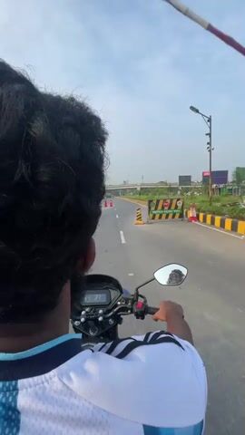 The video shows a person riding a motorcycle from a first-person perspective. The rider is wearing a white t-shirt with blue accents. The motorcycle's digital display shows a speed of 33. The road is paved and has a yellow and black striped barrier on the right side. There are orange traffic cones on the road ahead. In the distance, there is an overpass and some buildings. The sky is cloudy.