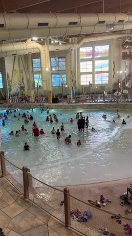 The video shows an indoor water park with a large wave pool filled with people of various ages, including children and adults, enjoying the water. The pool has gentle waves. In the background, there are water slides and other attractions. The environment appears to be a recreational facility, with artificial trees and decorative elements contributing to a themed atmosphere. People are swimming, playing, and relaxing in and around the pool. Several lounge chairs and towels are visible near the edge of the pool.