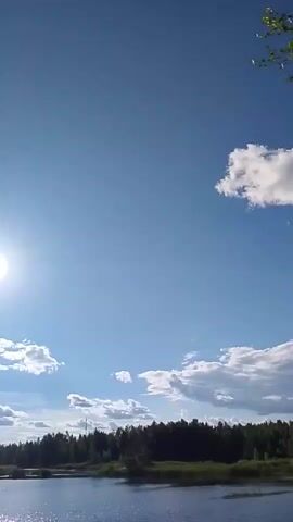 This is a vertical, stabilized video of a daytime outdoor scene. The sky is bright blue with scattered white cumulus clouds. On the left side of the frame, a bright sun is visible, causing lens flare. In the center of the frame, slightly above the sun, is a smaller, faint white light. Below the sky, a dark line of trees forms the horizon. In the foreground, a body of water reflects the sunlight, creating a shimmering effect. The water appears calm. There are no people or animals visible in the video. The video appears to be filmed from a stationary position.