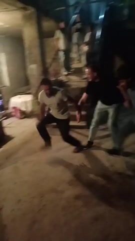 In a dimly lit, outdoor courtyard area that appears to be in Aragatsavan, Armenia, a group of people are engaged in a lively celebration. The scene is somewhat rustic, with concrete flooring and stone-like walls visible. In the foreground, three men are actively dancing, their movements energetic and coordinated. One man, wearing a light-colored t-shirt and dark pants, is at the center of the activity, his arms outstretched and body in motion. To his right, a man in a black t-shirt and light-colored pants is also dancing, his expression seemingly engaged and joyful. To the left, another man in a dark t-shirt and pants participates in the dance, his posture dynamic.

Behind them, a set of stairs leads up to an area where several other individuals are observing the dancing. Some are standing, while others are seated, suggesting a communal gathering. The lighting suggests it is either late evening or early night. The overall atmosphere appears to be one of festivity and shared enjoyment, with the dancing being the central activity. There is no visible text in the media.