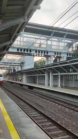 A train approaches and passes a station platform. The train is white with red accents and features the "KAI" logo. The platform has a yellow safety line and a covered waiting area with white pillars. An overhead pedestrian walkway with glass railings is visible in the background, and trees can be seen beyond the station. The sky is overcast.