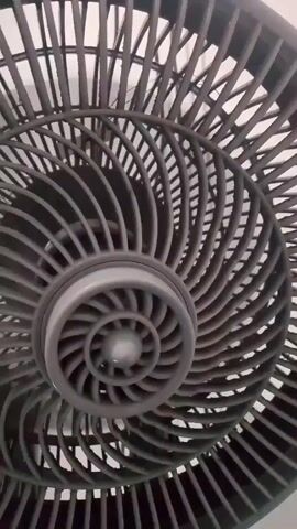 The video shows a close-up shot of a dark grey electric fan with its blades spinning. The camera is positioned directly in front of the fan, capturing the intricate design of the protective grill and the motion of the blades. The fan appears to be stationary, and the lighting suggests an indoor environment. The focus is solely on the fan and its operation.