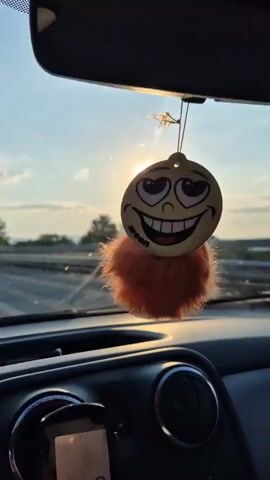 This media file captures a view from inside a car, looking forward. The primary subject is a dangling air freshener shaped like a smiley face with large eyes and a wide grin, attached to the rearview mirror. It has a fluffy orange pom-pom at the bottom. The sun is setting or rising, casting a warm, bright glow through the windshield. The light creates lens flare and illuminates the car's dashboard, which includes a phone mount and air vents. The background shows a road, trees, and a hazy sky, suggesting the car is in motion. The overall mood is cheerful and relaxed, conveyed by the smiley face and the warm lighting. The visible text on the air freshener reads "areon". The location is identified as Osikovitsa, Bulgaria, implying the scene is set in a rural or semi-rural area of the country.