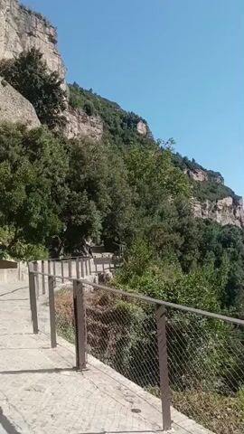 A video pans up a sheer cliff face on a sunny day. A paved path with a metal railing runs along the base of the cliff. Trees and vegetation grow on the cliff and surrounding slopes. The sky is clear and blue.