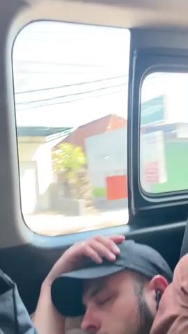 A man sleeps in a moving vehicle. He wears a brown t-shirt, black baseball cap, and small earbuds. He rests his head on a pillow that reads "All About Vacation". The camera pans out slowly showing the vehicle interior including a backpack and a window showing trees passing by.
