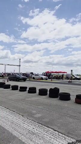 A dark sedan is shown on a drag strip, accelerating from a standstill. The car begins to move forward, and a person in the foreground is watching the event. The sky is blue with scattered clouds.
