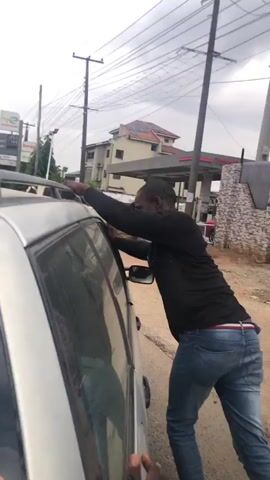 The video shows a man in a black long-sleeved shirt, blue jeans, and a black cap leaning on the roof of a silver car. His right hand is on the roof rack of the car, and his left hand is resting on the side of the car. He appears to be pushing himself up or trying to get into the car. His left leg is bent, and his right leg is extended. Two hands are visible in the foreground, one on the side of the car near the man's left hand, and another below it. The background shows a street with a gas station, buildings, power lines, and other cars. The ground is dirt and gravel. The overall scene suggests an informal or possibly tense interaction involving the man and the car.