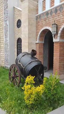 A slow pan reveals a courtyard in front of a stone building with ornate architectural details. On the right, a large, dark barrel on wooden wheels sits in a patch of grass with yellow flowers. To the left of the barrel, several large, earthenware amphorae are nestled in the grass. In the background, a building with two towers and arched windows dominates the view. The courtyard is paved with gray stones. The sky above is cloudy.