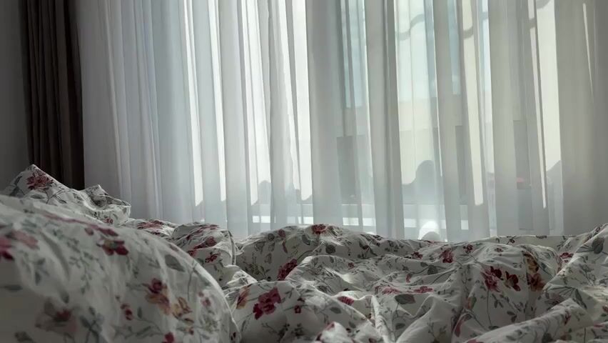 A close-up shot of a rumpled bed with floral patterned sheets. Sheer white curtains are visible in the background, with sunlight filtering through them. The scene is quiet and still.