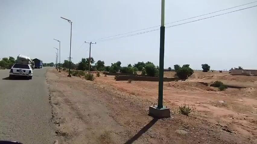On a sunny day, the camera films from the back of a moving vehicle, driving on an asphalt road. Ahead, the road leads past sporadic trees and a car with a heavy load strapped to its roof, followed by a semi-truck. The roadside is a dry, reddish soil interspersed with sparse vegetation, and lined with evenly spaced streetlights and telephone poles.