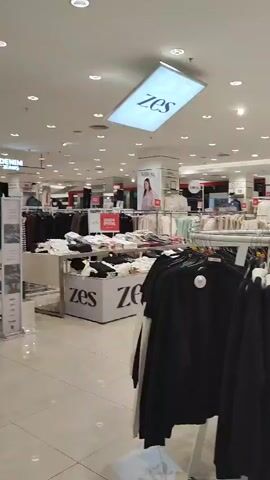 The video shows an interior view of a clothing store. Racks of clothing are displayed throughout the store, with items such as jackets, sweaters, and pants visible. The store appears to be well-lit with overhead lighting. Several displays and signs are present, some of which contain brand names or sale information. In the background, mannequins are used to display clothing. The overall environment suggests a retail setting focused on apparel.
