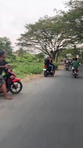 The video shows a group of people riding motorcycles on a paved road. In the foreground, two individuals are riding on a black motorcycle. The person in the back is looking over their left shoulder. The rider is wearing a black t-shirt and a white cap. The passenger is wearing a black t-shirt. To the right of the foreground motorcycle, there are three other motorcycles in the distance, also carrying two people each. The road is lined with lush green vegetation on the left and trees on the right. The sky is overcast.