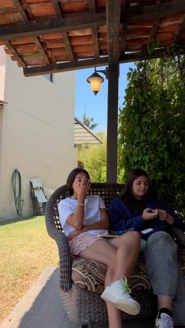 Here is a concise description of the video:

The video opens with a group of teenagers sitting and interacting in an outdoor covered patio area. They are sitting on benches and rattan furniture around a wooden table. The camera pans to show another group of teens sitting nearby. The camera then moves to a scene of a dog walking along the edge of a pool. In the background, there is a chain-link fence, rolling hills, and a playset. The sky is blue with a few clouds.