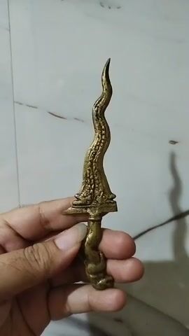 A hand is holding a miniature, ornate, wavy dagger. The dagger appears to be made of brass or a similar metal and has intricate detailing along the blade and handle. The background is a pale, marbled surface, likely a countertop or floor. The video shows the dagger from slightly different angles as it is rotated in the hand.