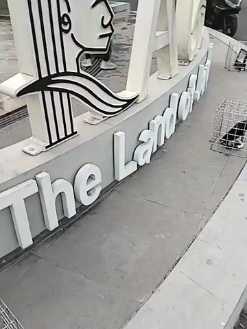 The video shows a statue display in a park. The first shot displays a white sign that reads "ACEH The Land of Java." The letter "A" in "ACEH" is a stylized profile of a person's head and neck. The next shot shows the same sign in a wider view. In the background, there is a large statue of a muscular human-like figure, a fountain with water jets, and a structure resembling large tusks. Several people are visible in the background, some walking and others playing in the fountain. The sky is overcast.