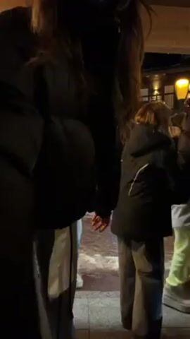 A group of people stand outside at night. The focus is on a young woman in a black puffer jacket, gray pants, and a black beanie, with a small crossbody bag. She is standing on one leg and making a peace sign with her hand. Other people are visible around her. The background includes a building with a lit window and an overhanging roof.