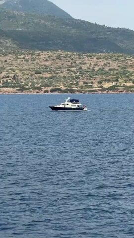 The video shows a yacht moving across the water near a coast with hills and light vegetation. The sky is partially cloudy and the water is blue with some ripples. The yacht is dark-colored with white accents. The camera appears to be on another boat moving through the water.