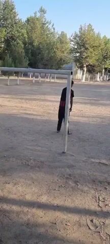 A person in a black shirt with red stripes on the sleeves and black pants is standing near a vertical pole in an outdoor area. The ground is dirt and gravel. In the background, there are trees and buildings. There are also several other people in the distance. The person in the foreground turns and walks away from the pole.