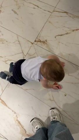 A baby lies prone on a glossy, light-colored, marble-patterned tile floor, attempting to crawl. The baby is wearing dark pants and a light blue or white shirt and patterned blue socks. Directly in front of the baby's head, a person wearing blue jeans and silver and white sneakers stands stationary, providing a visual reference point. The camera angle is directly overhead, looking down at the scene.