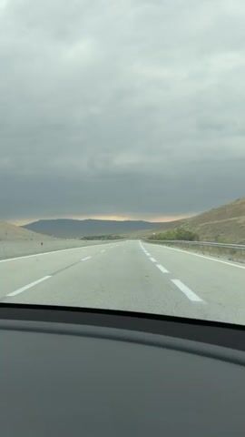 The video shows a view from a car on a highway. The sky is overcast with heavy, gray clouds. The road ahead is empty and asphalt, with white lane markings. To the right of the road, there is a metal guardrail and a dry, hilly landscape with sparse vegetation. In the distance, a range of hills or mountains is visible. The lighting suggests it is either early morning or late afternoon.