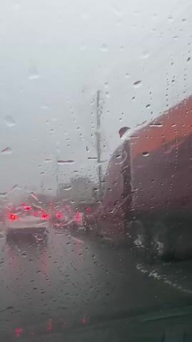 The video shows a view from inside a vehicle looking out through a rain-streaked windshield. On the road ahead, traffic is visible, with the red taillights of cars illuminating the wet pavement. A large, dark red truck with an orange section is on the right side of the frame, partially obscured by the raindrops on the glass. The overall scene is grey and overcast, indicating poor weather conditions.