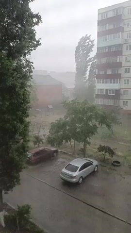 The video shows a courtyard during a heavy storm. The sky is overcast and gray, indicating a significant amount of precipitation. The wind is strong, as evidenced by the trees being blown severely to the side. Rain is falling in sheets, making it difficult to see distant objects clearly. There are two cars parked in the courtyard, a red station wagon and a silver sedan. One car appears to be older and possibly worn, while the other is newer. In the background, there is a playground with swings and a slide, suggesting this is a residential area. A tall apartment building is visible on the right side of the frame, with multiple stories and windows. The overall impression is one of a severe weather event impacting an urban or suburban environment.